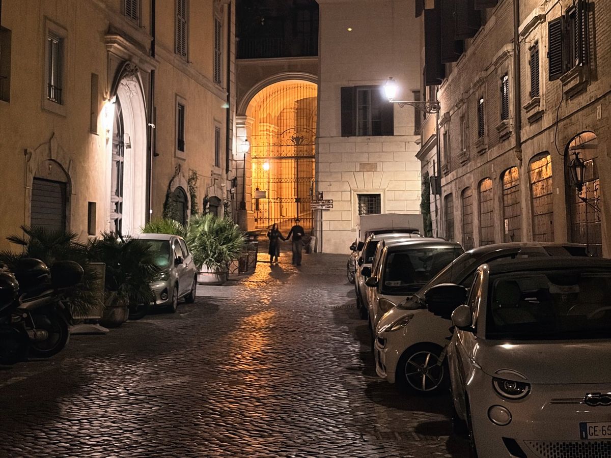 Narrow cobblestone street at night with parked cars and motorbikes, flanked by tall, shuttered buildings. A glowing archway illuminates two distant pedestrians under warm streetlights.