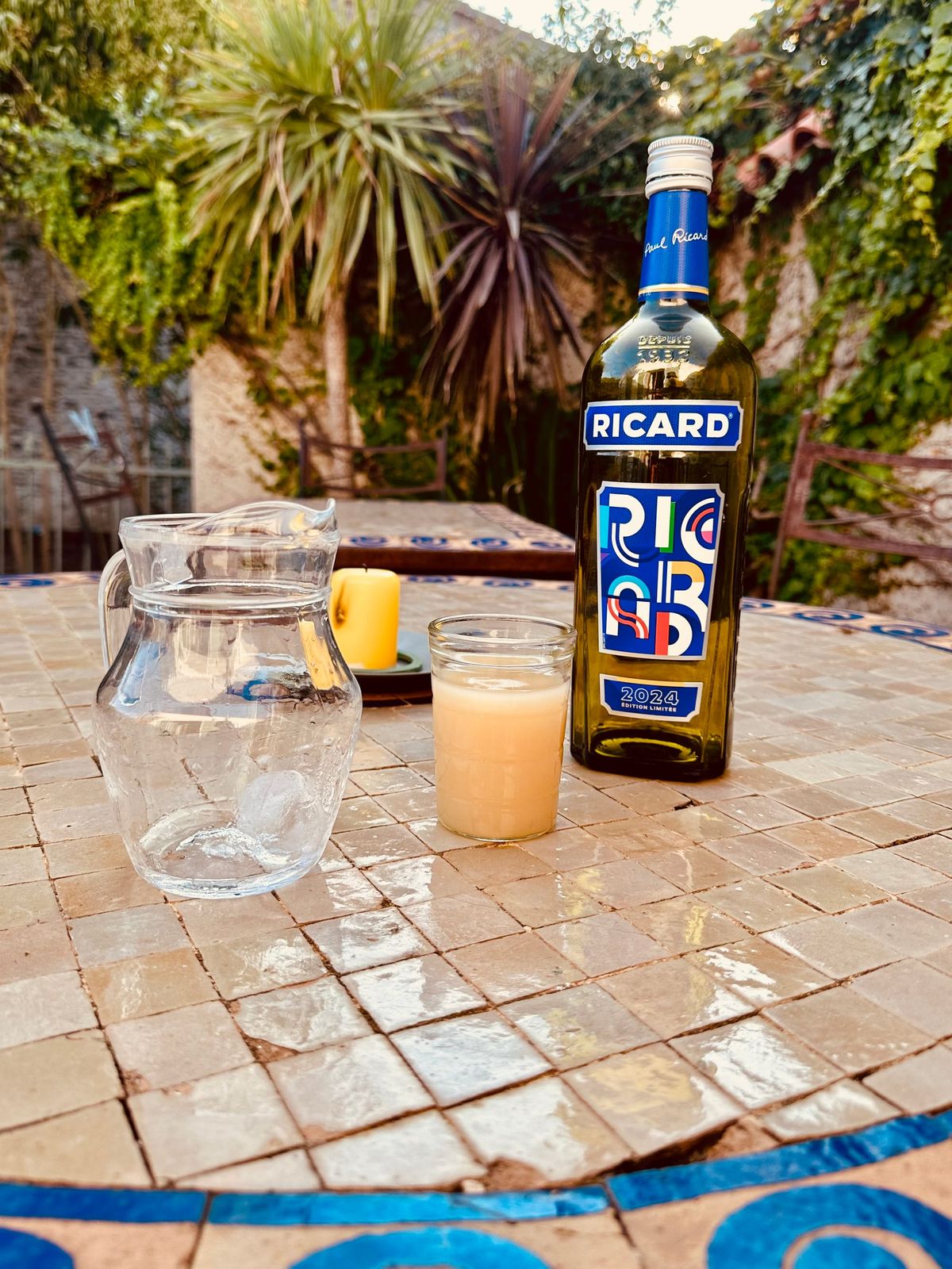 A tiled garden table on which are: a jug of water, a citronella candle, a glass of pastis, and a bottle of Ricard