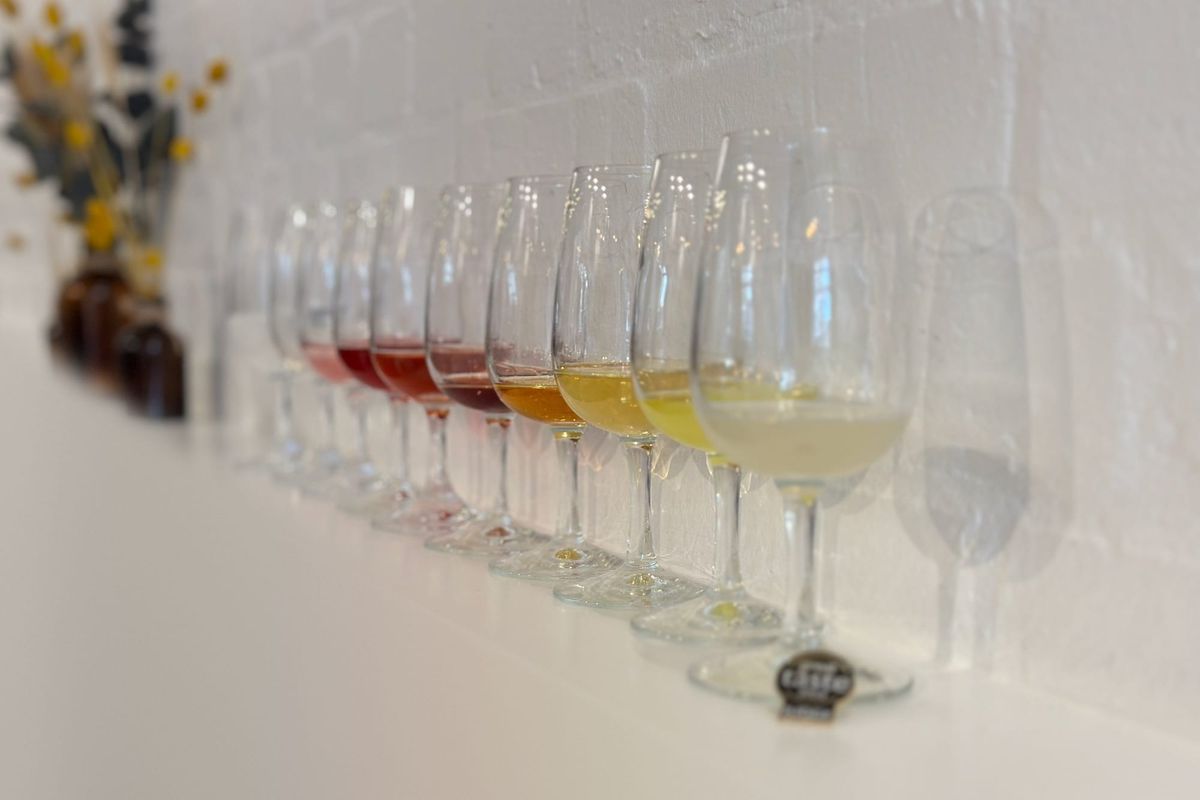 A row of tasting glasses on a long white shelf, shading from pale pink through deep red and amber to gold and almost clear, with dried flowers in the background.