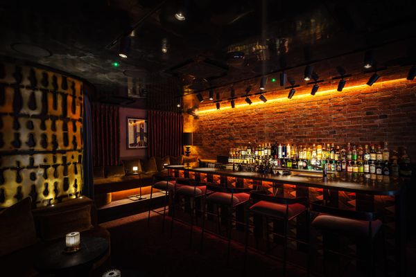 Warm, atmospheric basement cocktail bar with exposed brick walls, amber lighting, red bar stools, and backlit bottles behind the bar