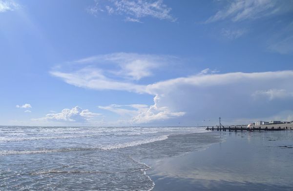 Calm waves lap against a wide sandy beach under a bright blue sky streaked with wispy and towering white clouds, with wooden groynes and small seaside buildings in the distance.
