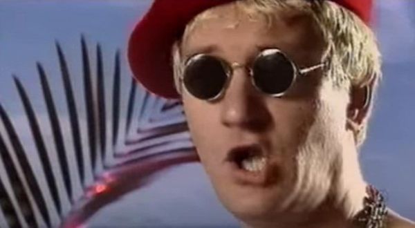 A man with short blond hair wears a bright red beret, round black sunglasses, and a chain necklace, singing with his mouth open against a sky-blue backdrop with a palm leaf.