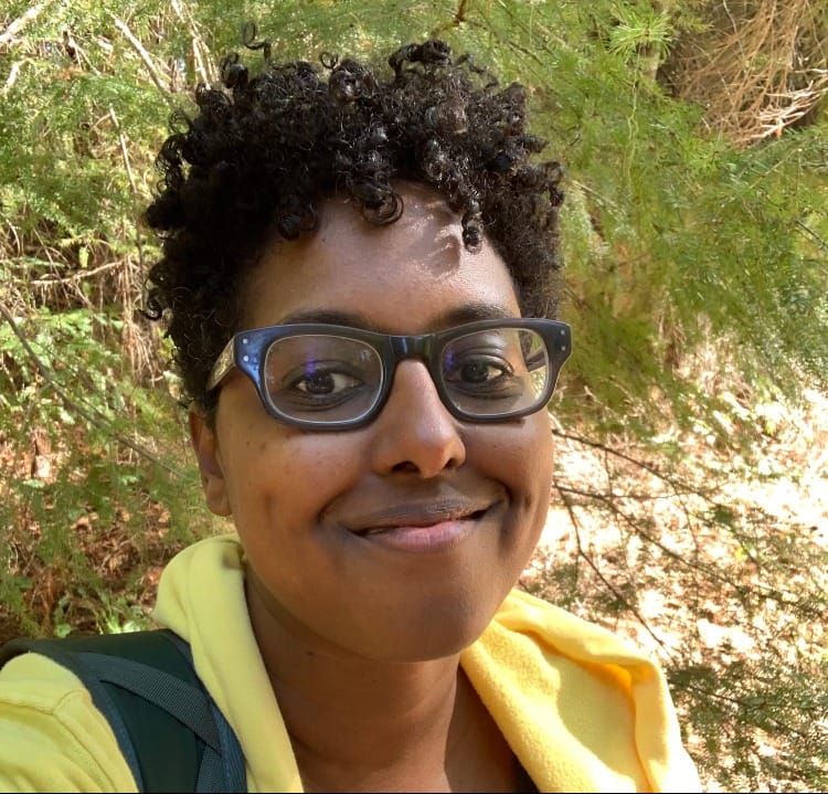 A close-up photo of a smiling black woman with glasses standing in front of trees.