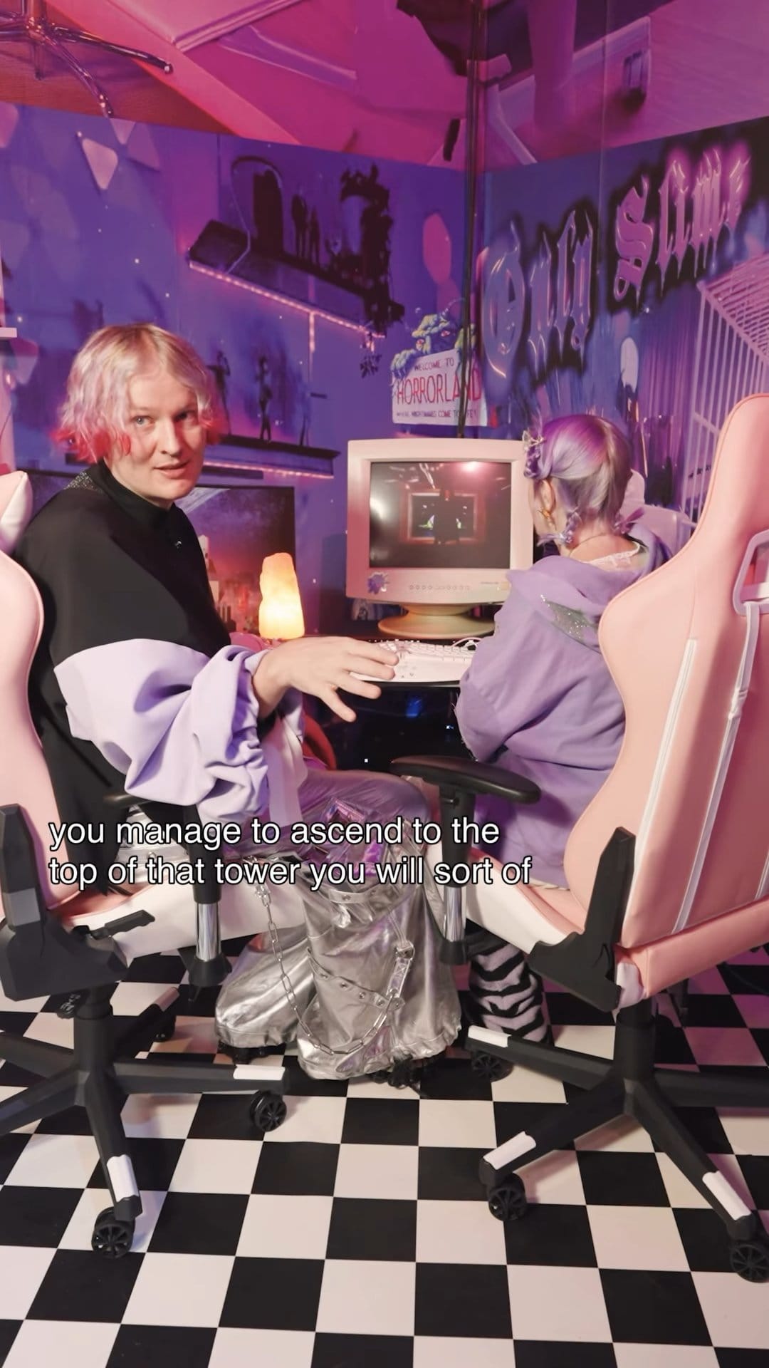 Two artists in costume at a CRT computer in pink gaming chairs on a checkered floor