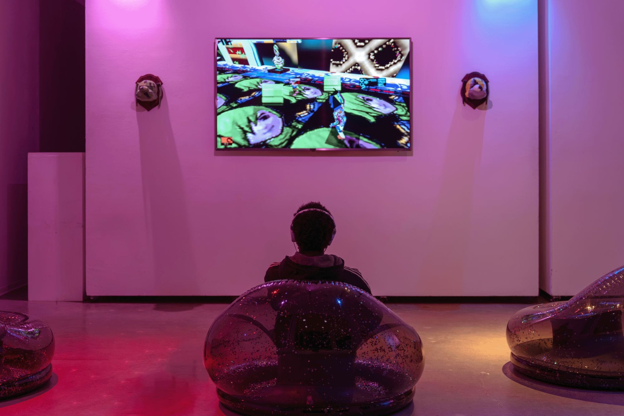 A visitor seated on an inflatable chair plays TEXTURETOWN on a wall-mounted screen in a pink-lit gallery