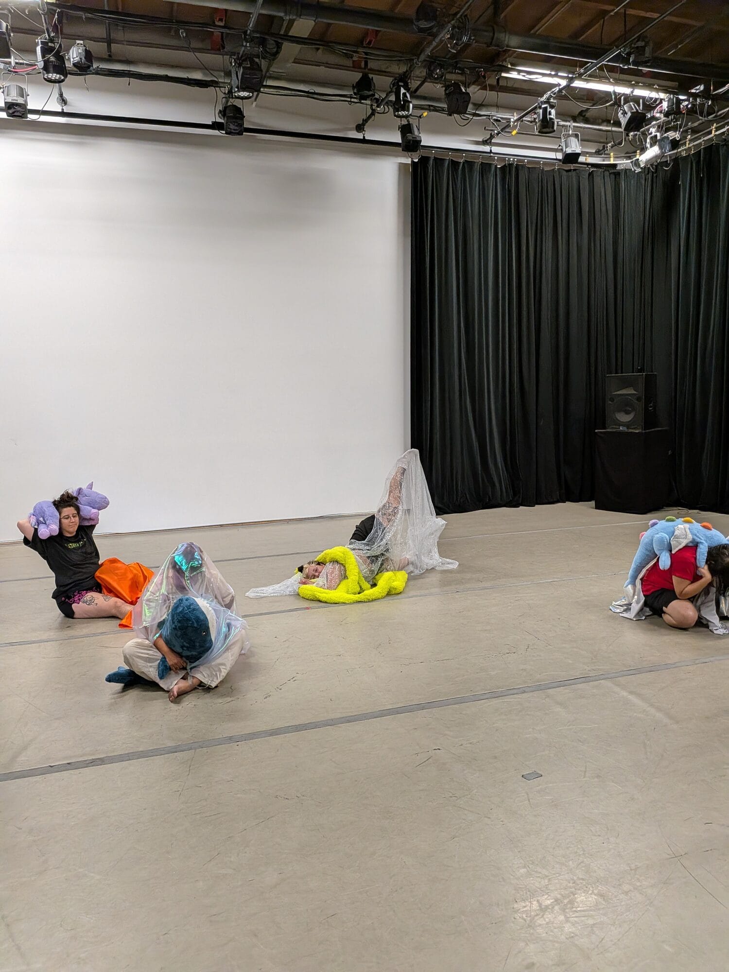 Performers collapsed on a rehearsal room floor wrapped in plush animals and cellophane