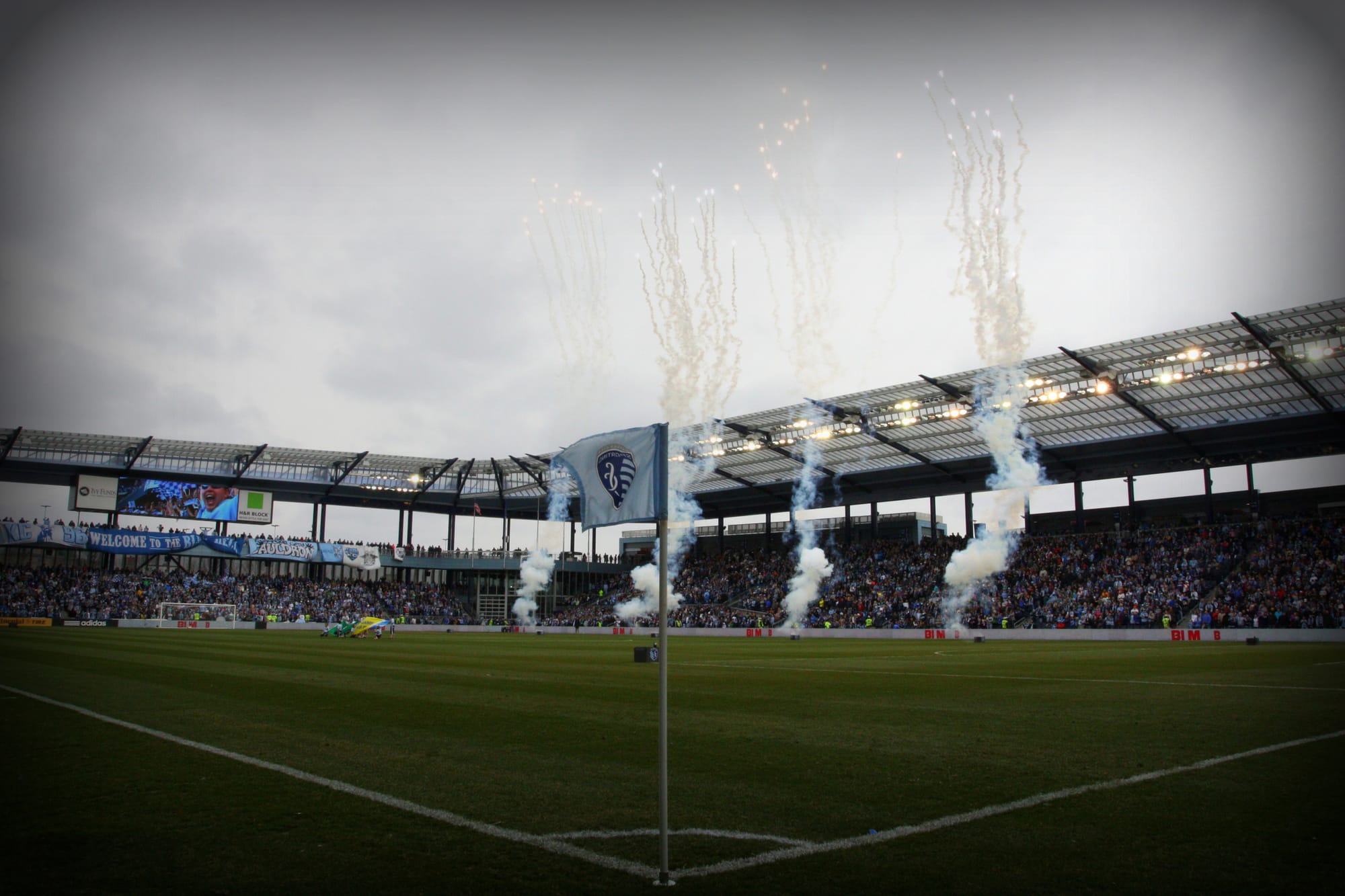 A floodlit soccer stadium at dusk, smoke plumes rising from the stands beyond the corner flag.