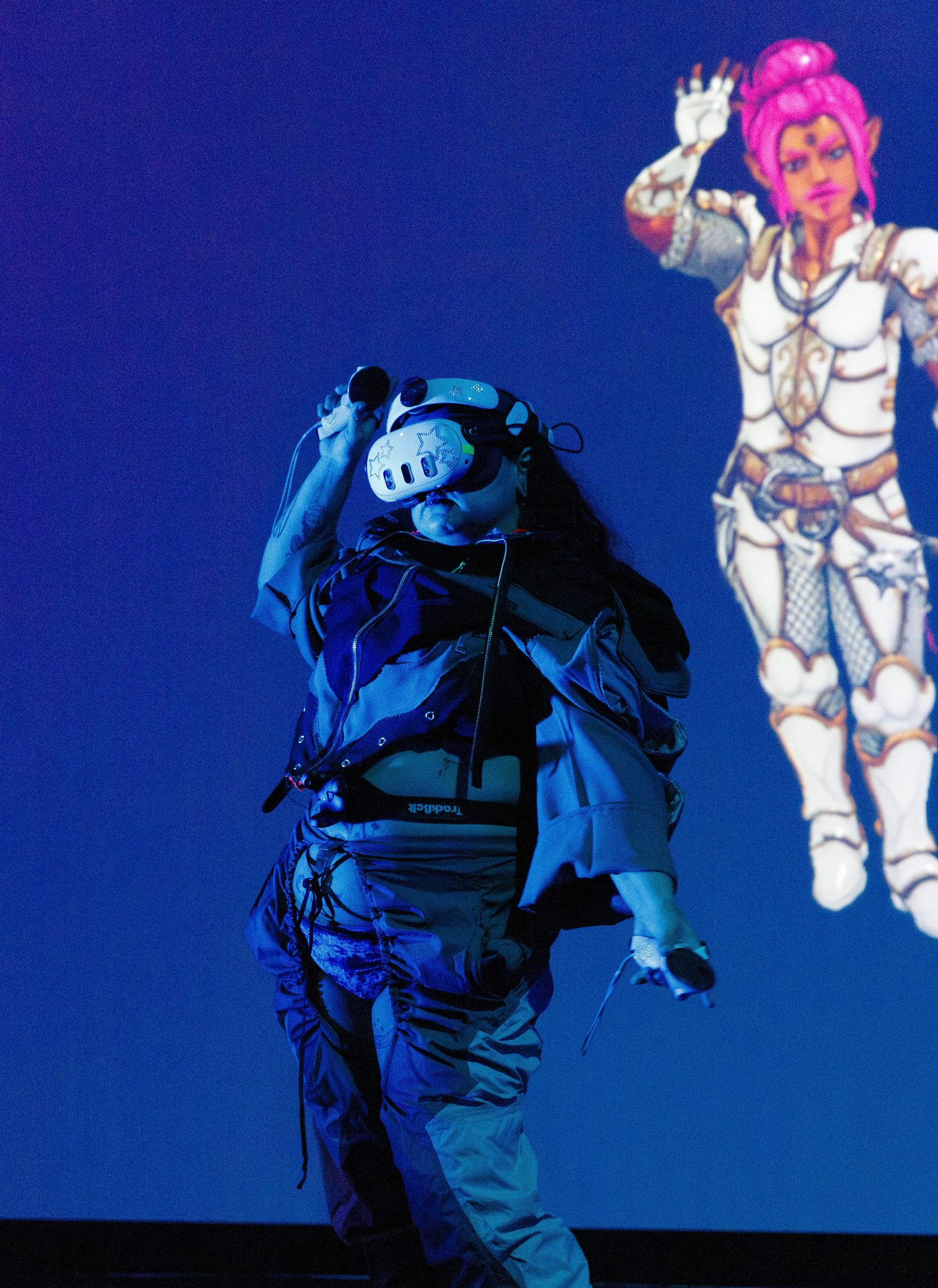 A performer in a full VR headset and motion capture rig raises one arm as a pink-haired armored avatar mirrors the pose on the screen behind