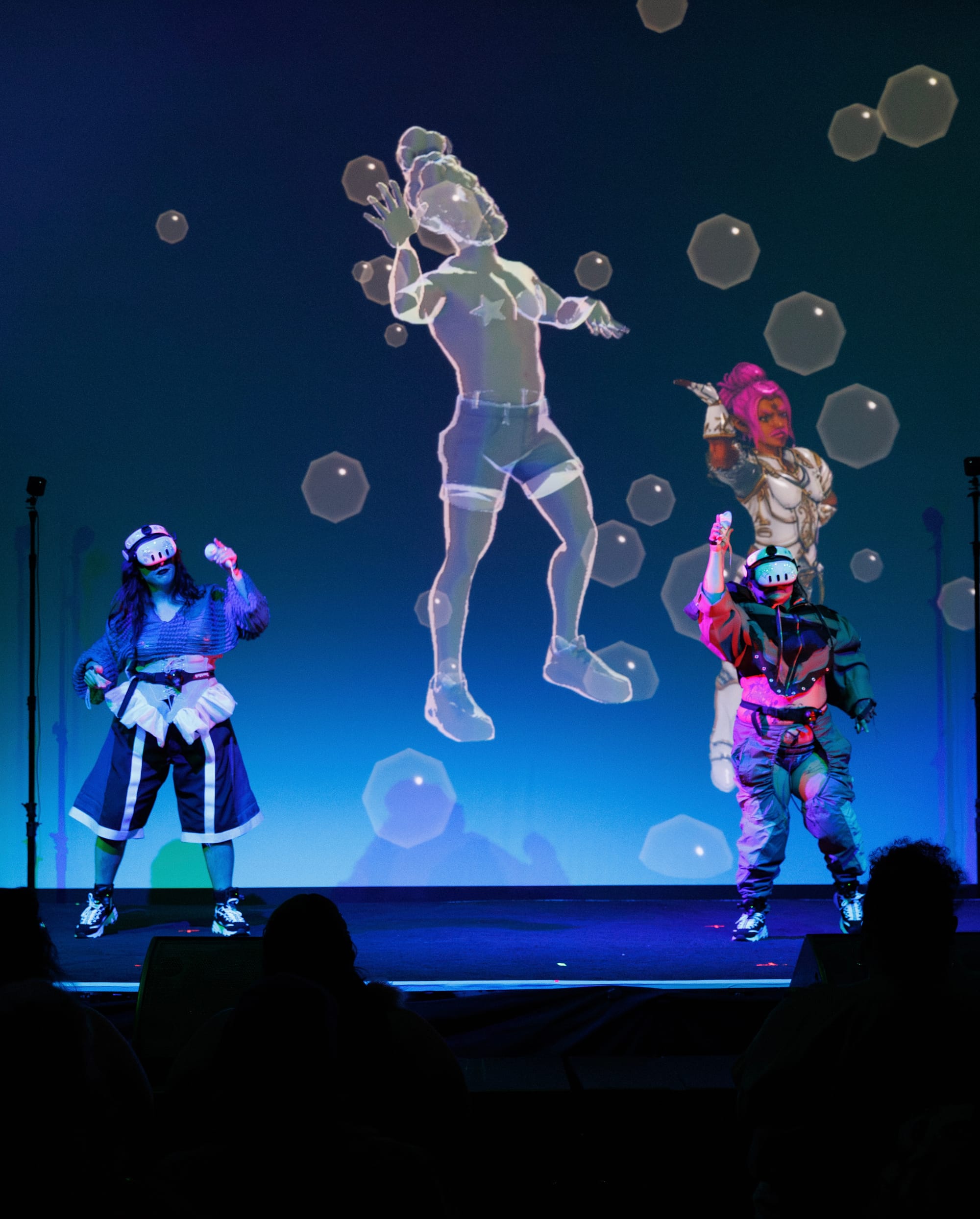 Performers in VR headsets beneath a large translucent avatar projected in blue light