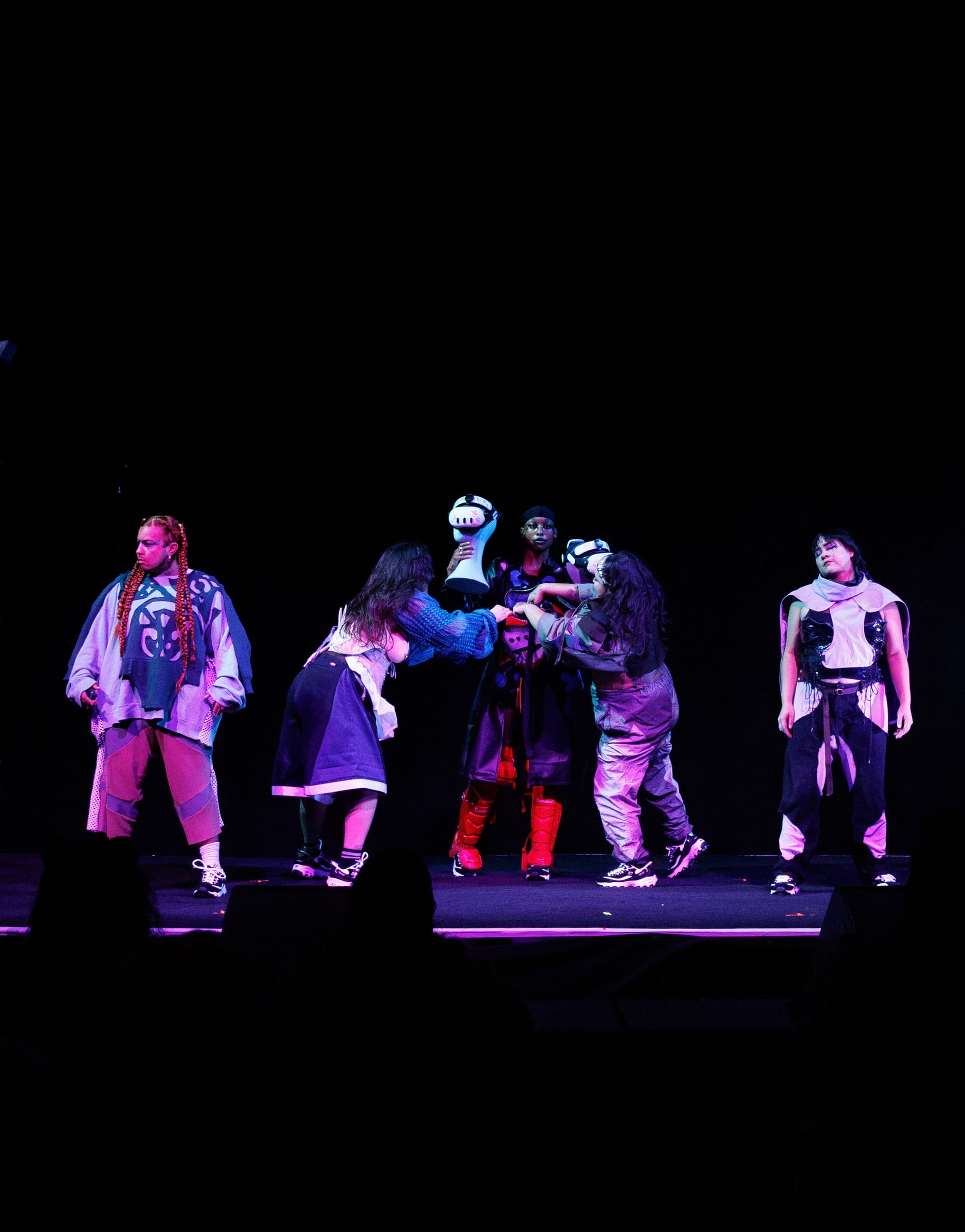 Performers clustered on a dark stage, one wearing a VR headset