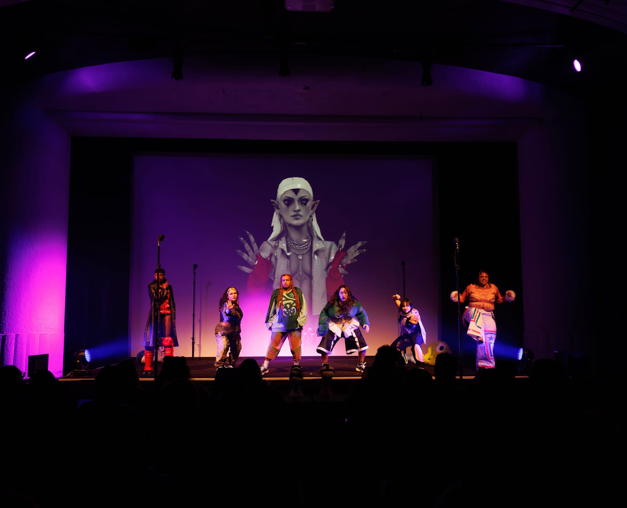 The full company onstage in front of a large projected avatar, audience silhouetted in the foreground
