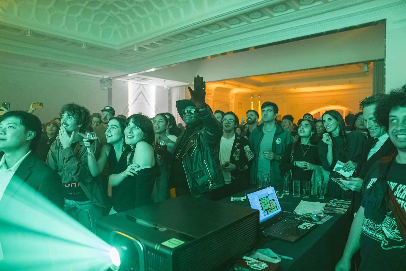 A crowd bathed in green projector light watches a screen at Rhizome's benefit afterparty