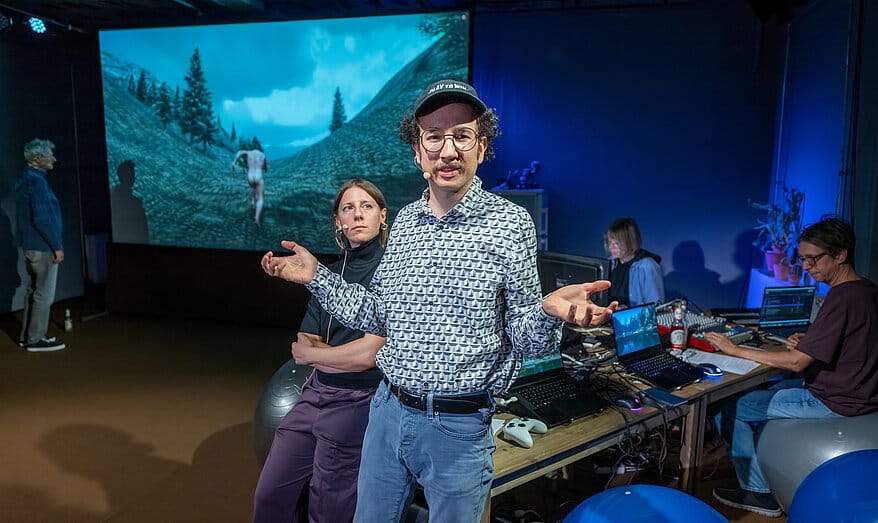 Two performers gesture in front of a projected forest scene while others work at laptops behind them