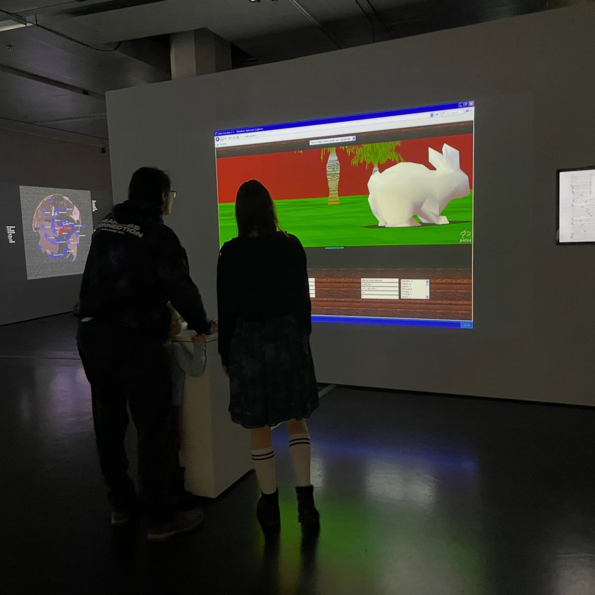 Two visitors watch a projection of Eden.Garden 1.0 showing a low-poly white rabbit in a virtual garden