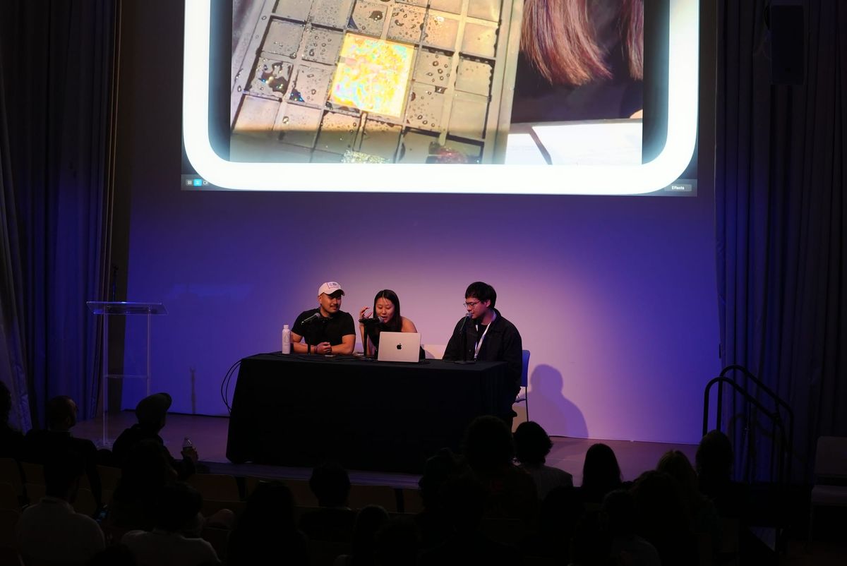 Three presenters at a table on a dark stage beneath a large projection screen at the New Museum