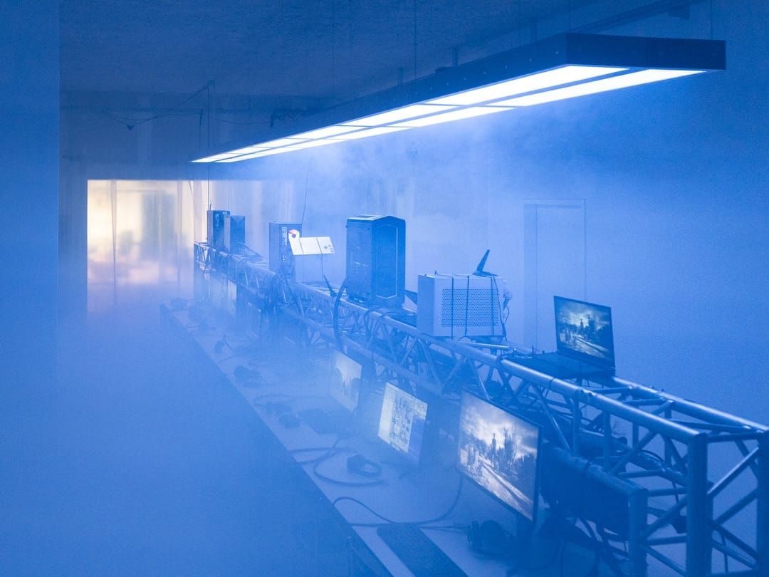 A row of gaming PCs and monitors on truss tables under blue light and fog at Sybil in Berlin