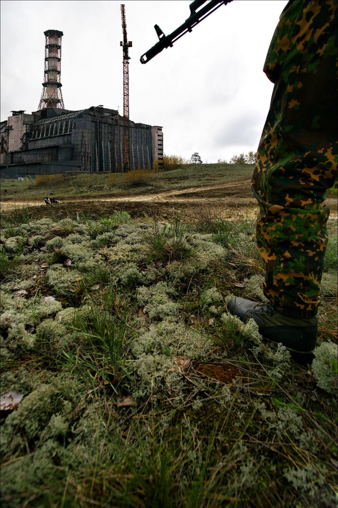The Chernobyl reactor building seen across an overgrown field, a figure in camouflage in the foreground.