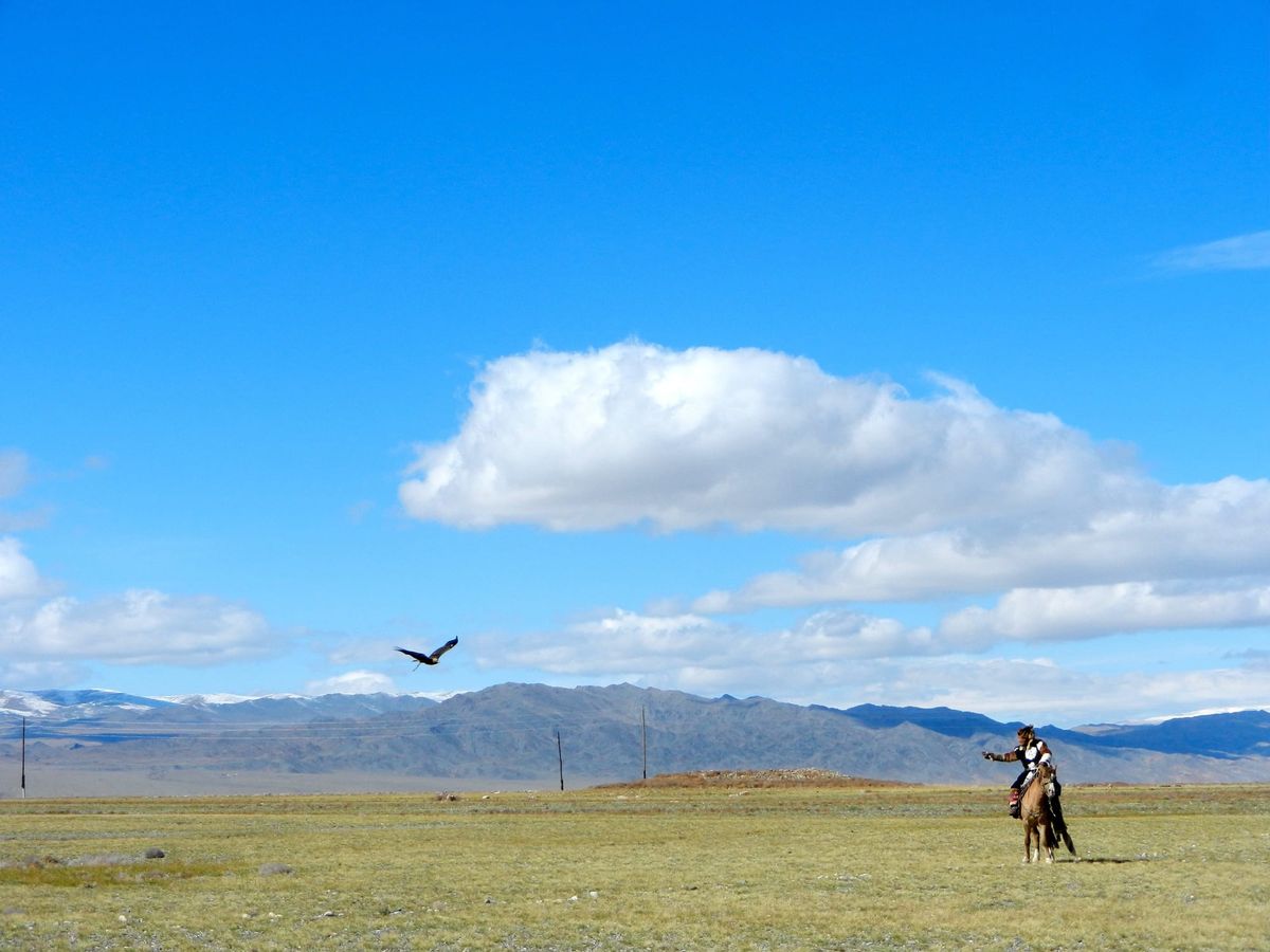 A lone horseman crosses an open Mongolian steppe under a wide blue sky as a bird circles overhead.