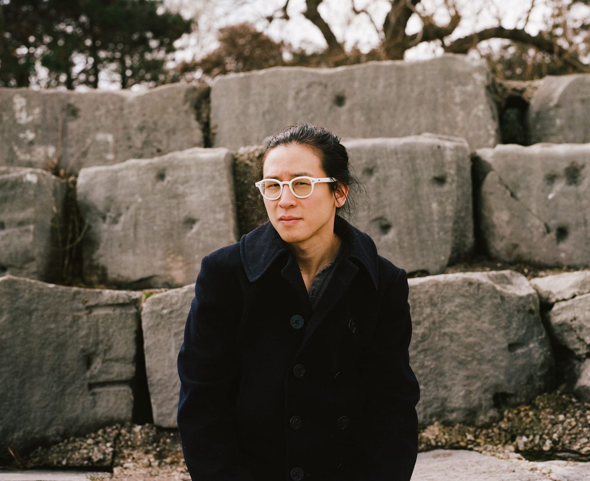 William Chyr in white-framed glasses and a dark coat, standing before a wall of stacked stone blocks