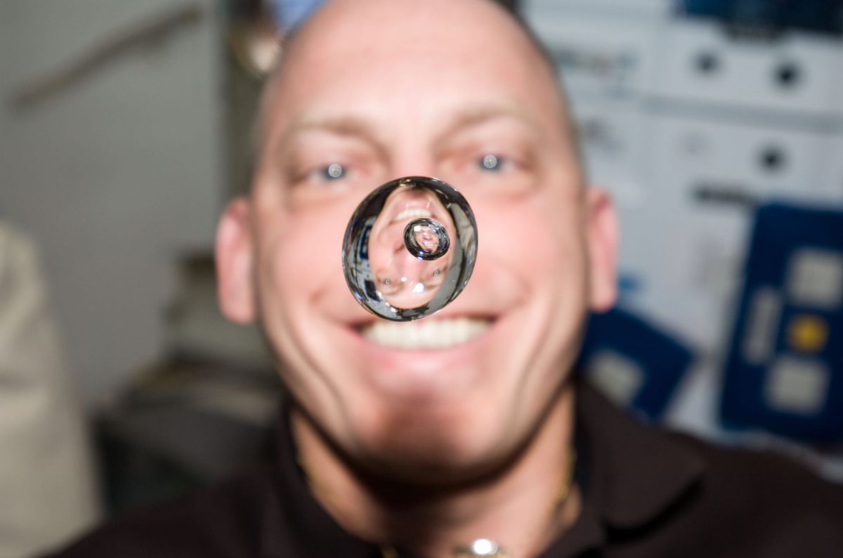 An astronaut aboard the space station grins behind a floating sphere of water that magnifies his face.