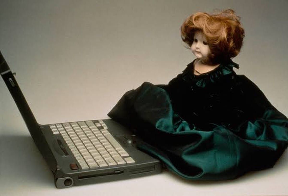 A doll in a dark green dress seated beside an open laptop, an image courtesy of Lynn Hershman Leeson