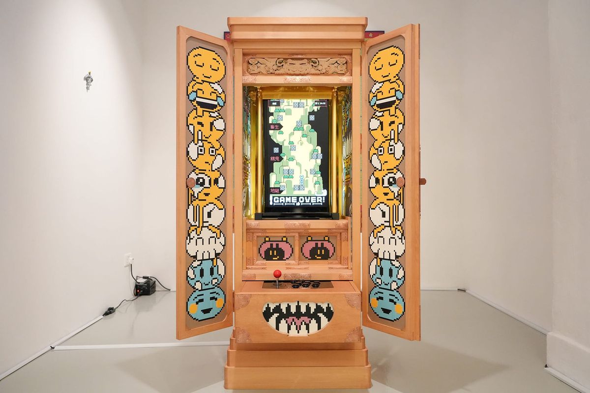 A wooden Buddhist household altar stands open in a white gallery, cartoon faces painted inside its doors and a vertical game screen glowing at its center.