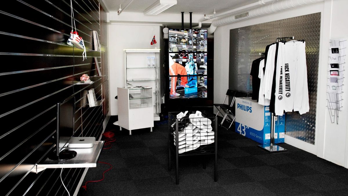 A small shop interior with black slatwall shelving, a Mac mini on a metal shelf, a wire bin of white pool slides, a video display and a rack of sweatpants.