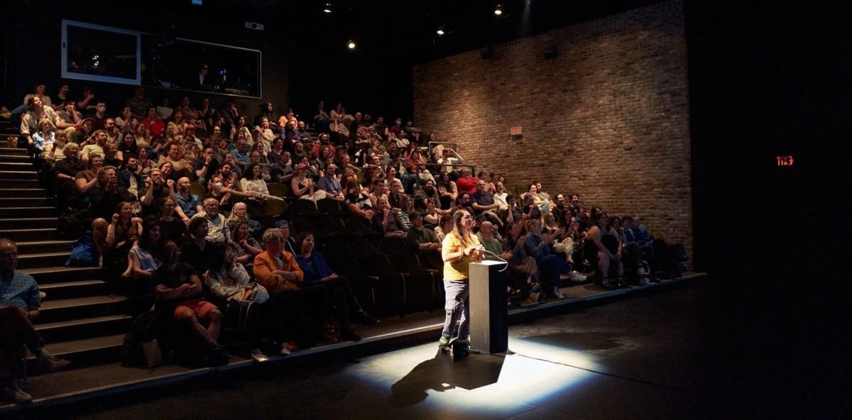 A full theater audience looks toward a screen while one person stands at a spotlit podium at the front of the room.
