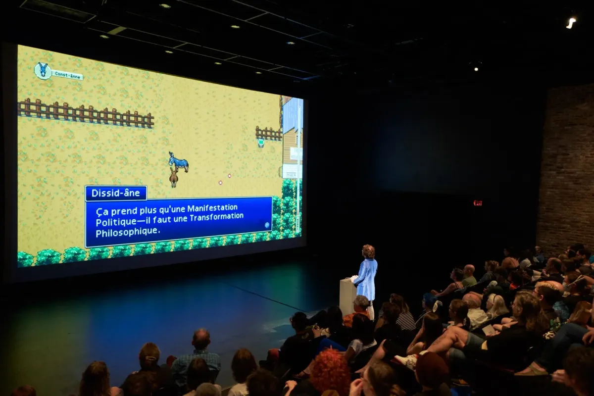 A theater audience in the dark faces a large screen showing the video game asses.masses