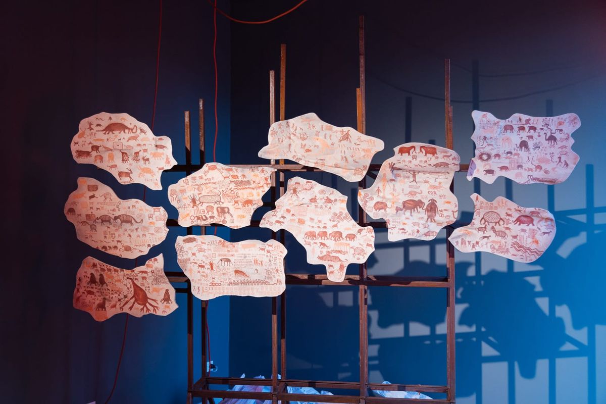 Ceramic plates painted with red and brown figures hang together on a metal frame in a dark blue room, lit by red lamps