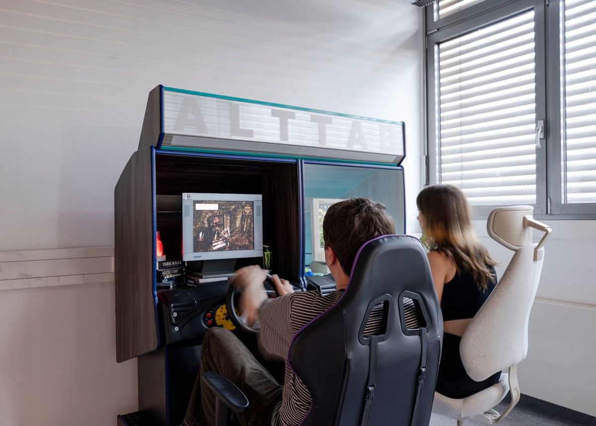 Two people sit at a pair of arcade racing cabinets labeled ALT TAB, each with a steering wheel and a screen.