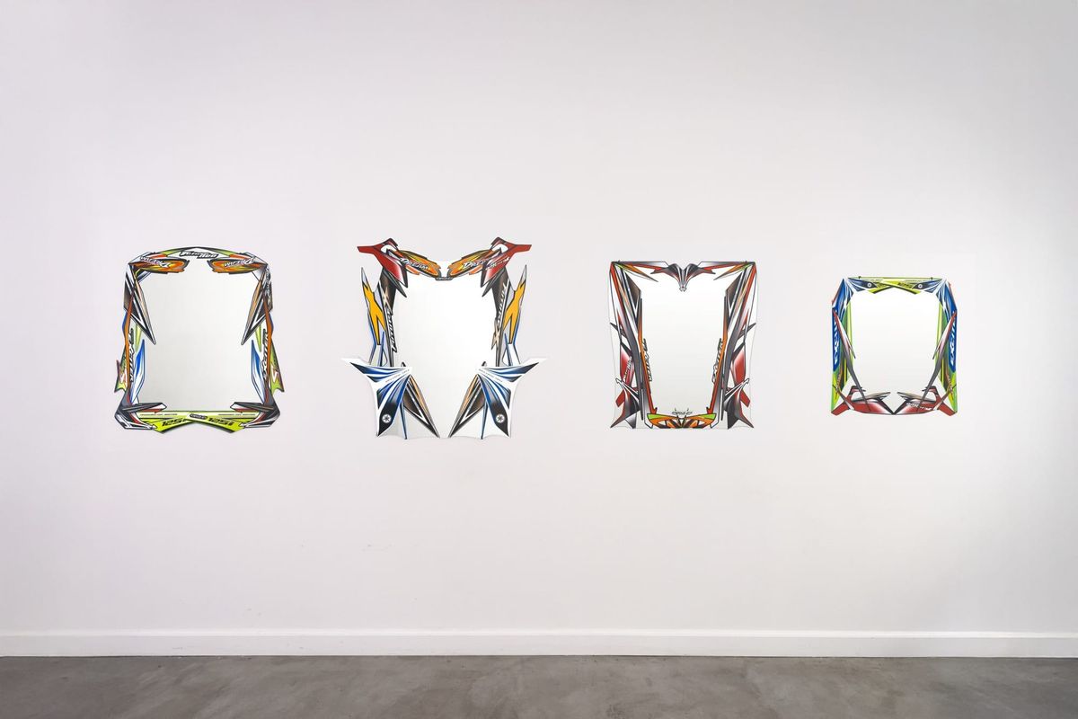 Four wall mirrors framed in colorful decal-covered scooter body panels, hung in a row on a white gallery wall.