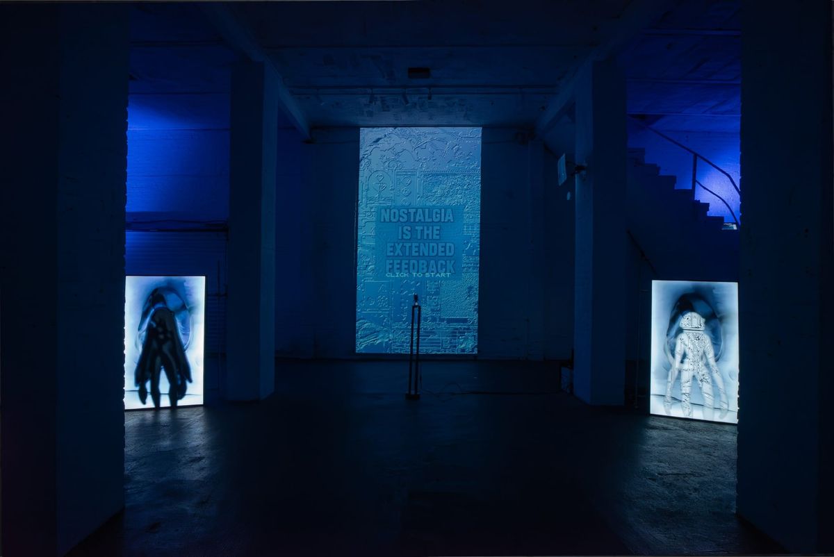 A dark gallery lit blue, with the words Nostalgia Is the Extended Feedback projected on the far wall between two upright glowing screens.