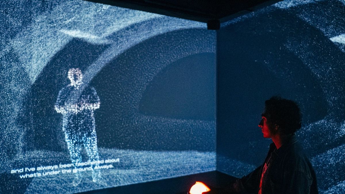 A visitor rests a hand on a glowing red orb in a dark room where a tunnel and a human figure are projected as clouds of blue-white points.