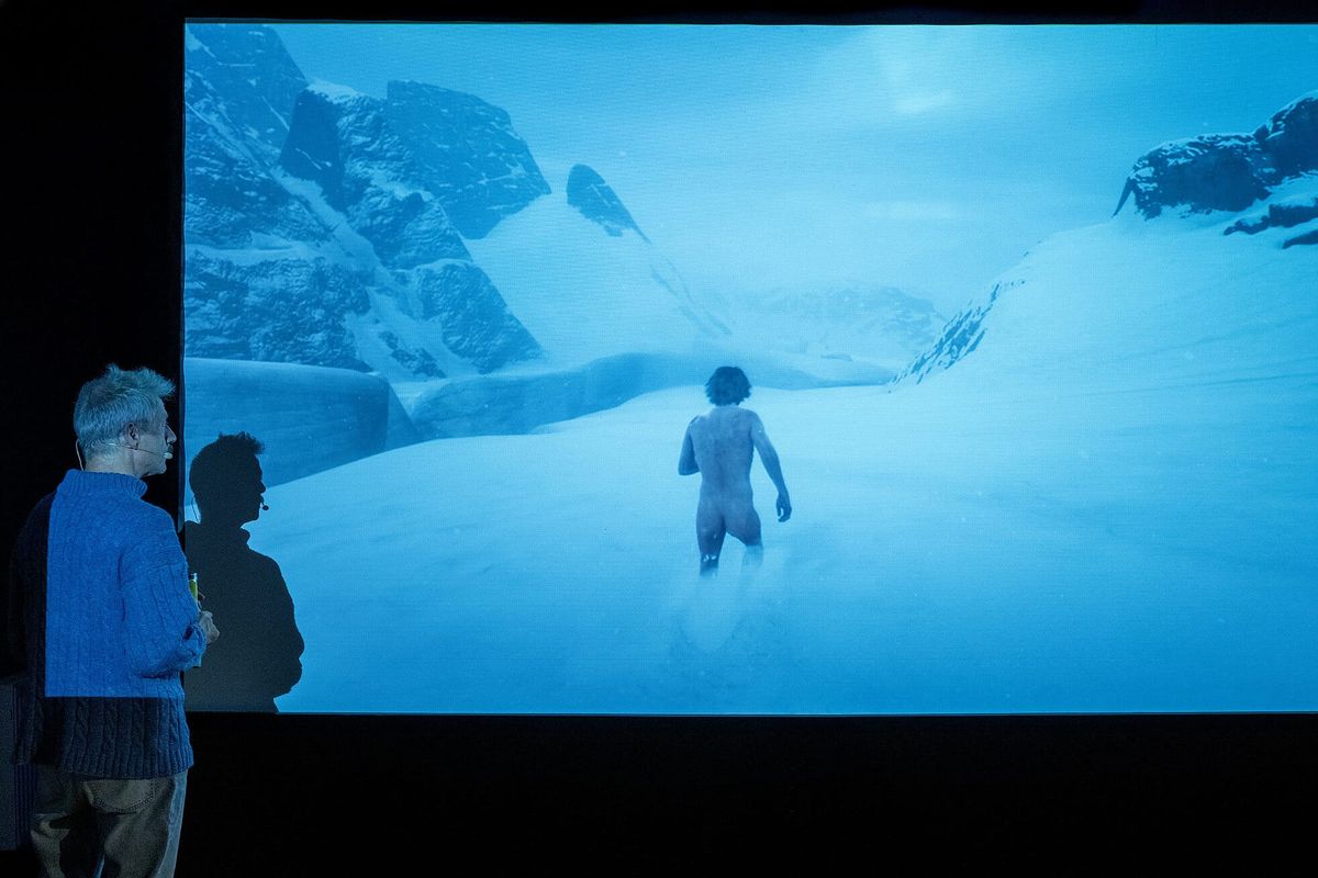 An audience in silhouette watches a projected game character wade through deep snow