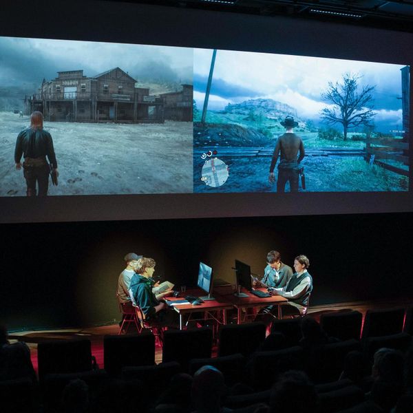 Performers seated at desks on a stage beneath two large projections of Red Dead Online, each showing a cowboy figure in a desert landscape