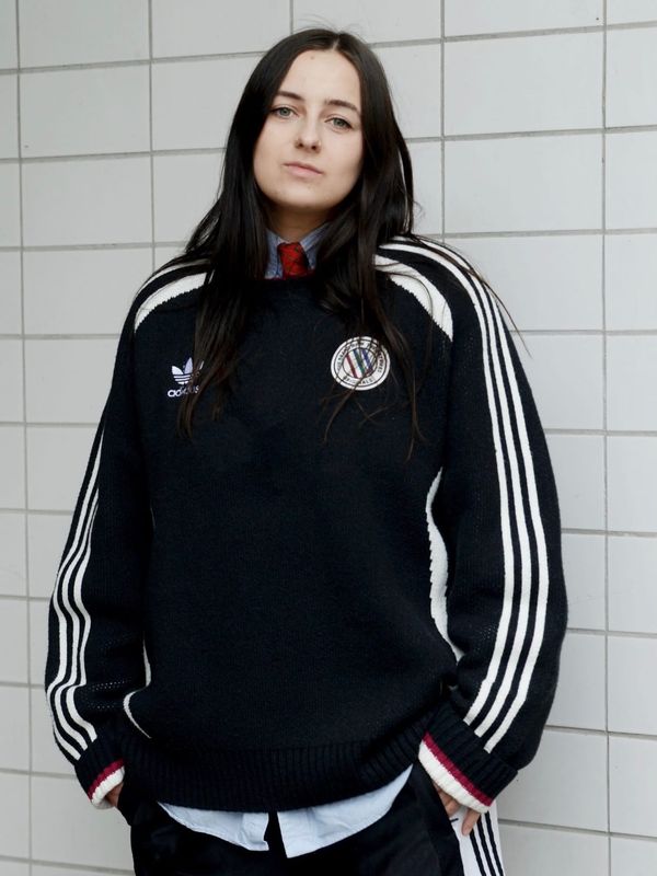 Lou Fauroux stands against a white tiled wall wearing a black track jacket.