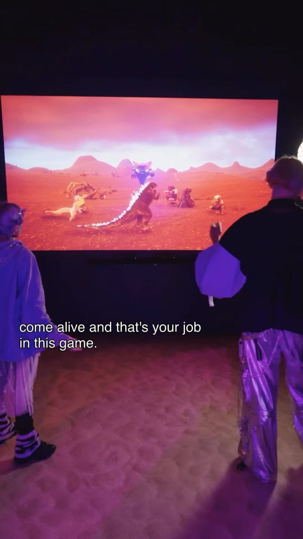 Two visitors stand before a large projection of a red desert game world with creatures