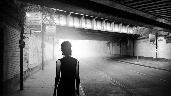 A silhouetted figure walks through a dark underpass toward bright light in a black-and-white scene from Signet City