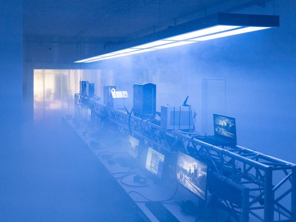 A row of gaming PCs and monitors on truss tables under blue light and fog at Sybil in Berlin