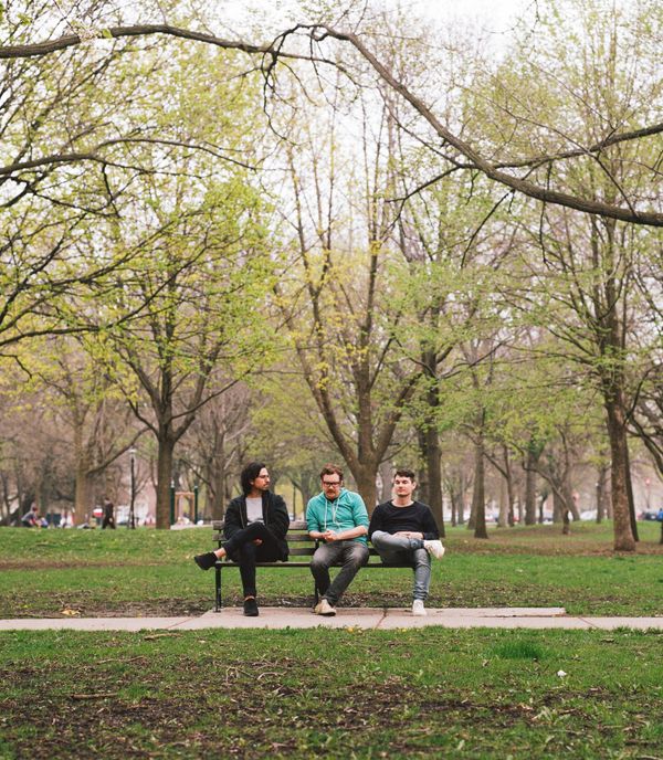 The three members of Cardboard Computer sitting on a park bench under spring trees