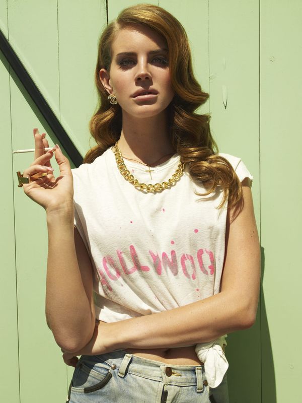 Lana Del Rey in a knotted white tee and gold chain, leaning against a pale green wall, cigarette in hand.