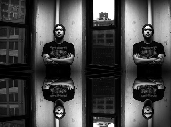 A black and white portrait of Mux Mool standing in a doorway, the frame tiled and mirrored into a repeating pattern.