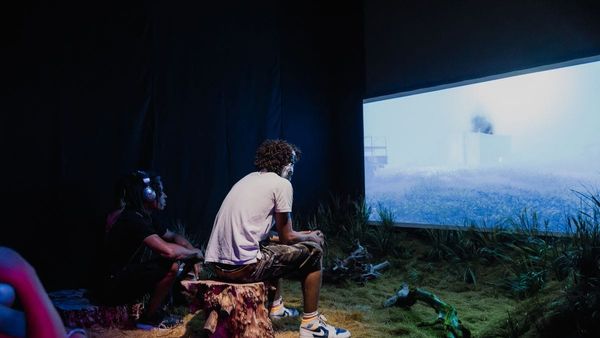 Visitors sit on tree stumps watching a large screen in the dimly lit Undershed gallery, with grass and branches on the floor