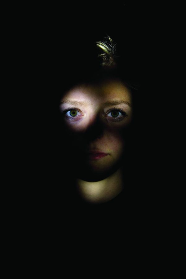 A face lit from below emerging out of near-total darkness, eyes open, the rest of the frame black.