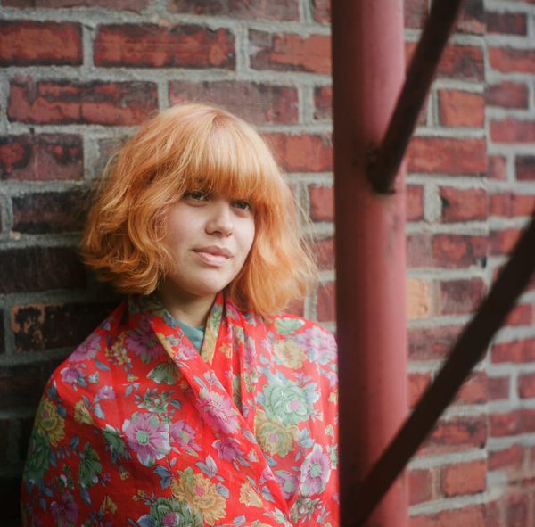 Paloma Dawkins in a red floral jacket beside a brick wall and painted red pipe