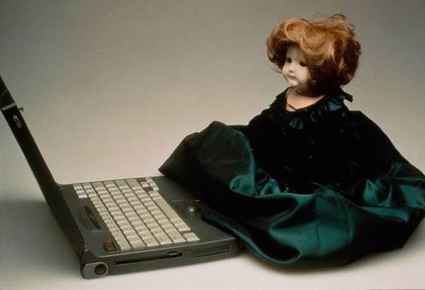 A doll in a dark green dress seated beside an open laptop, an image courtesy of Lynn Hershman Leeson