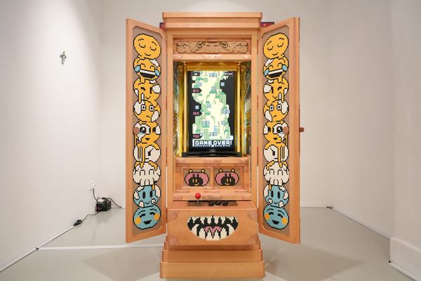 A wooden Buddhist household altar stands open in a white gallery, cartoon faces painted inside its doors and a vertical game screen glowing at its center.
