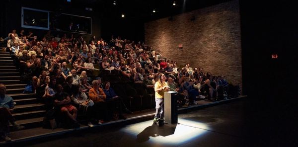 A full theater audience looks toward a screen while one person stands at a spotlit podium at the front of the room.