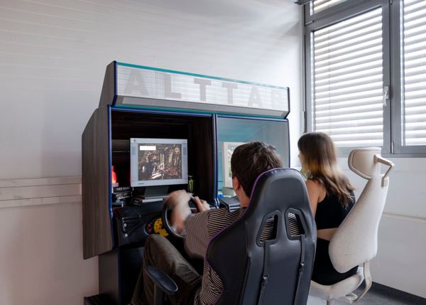 Two people sit at a pair of arcade racing cabinets labeled ALT TAB, each with a steering wheel and a screen.